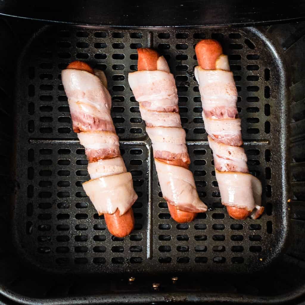hotdogs in air fryer basket wrapped in bacon and ready to be cooked