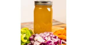 A mason jar filled with homemade Instant Pot vegetable broth, surrounded by chopped fresh vegetables including celery, red onion, and carrots, displayed on a wooden table.